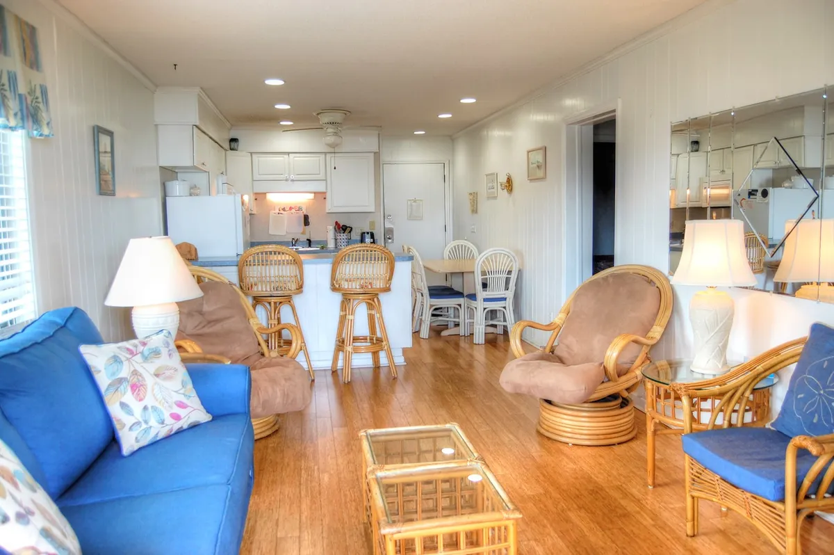 This image features a bright, open living room with a blue sofa, rattan chairs, a glass coffee table, and a partial view into the kitchen area. Large windows flood the room with natural light, highlighting the comfortable seating arrangement ideal for groups and families.