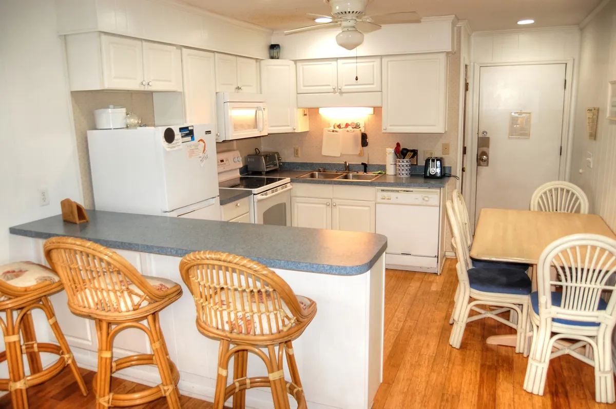 A cozy dining area with a kitchen island, bar stools, a dining table, and white cabinetry is shown. The scene is warm and welcoming, perfect for shared meals, game nights, or conversations over coffee.