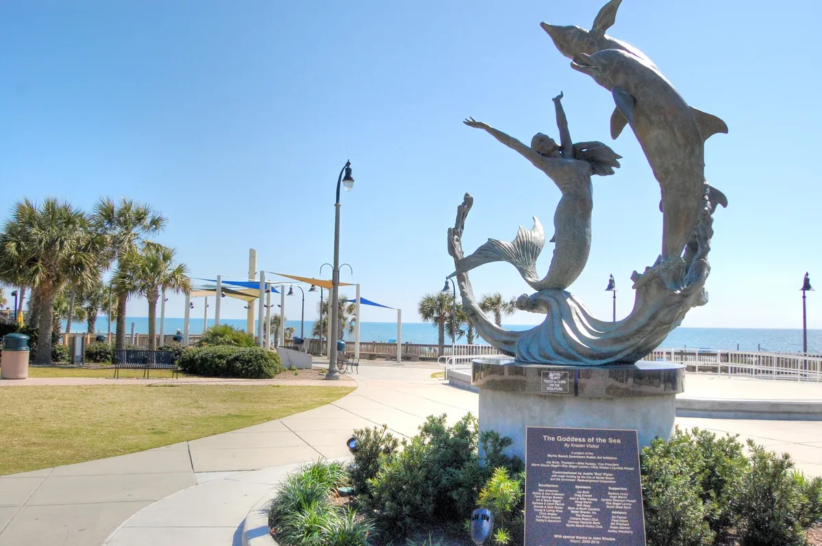 A scenic walkway lined with palm trees leads to a striking sculpture of the Goddess of the Sea, set against the ocean and open sky. The area invites leisurely strolls, group photos, and moments of togetherness in a beautiful setting.