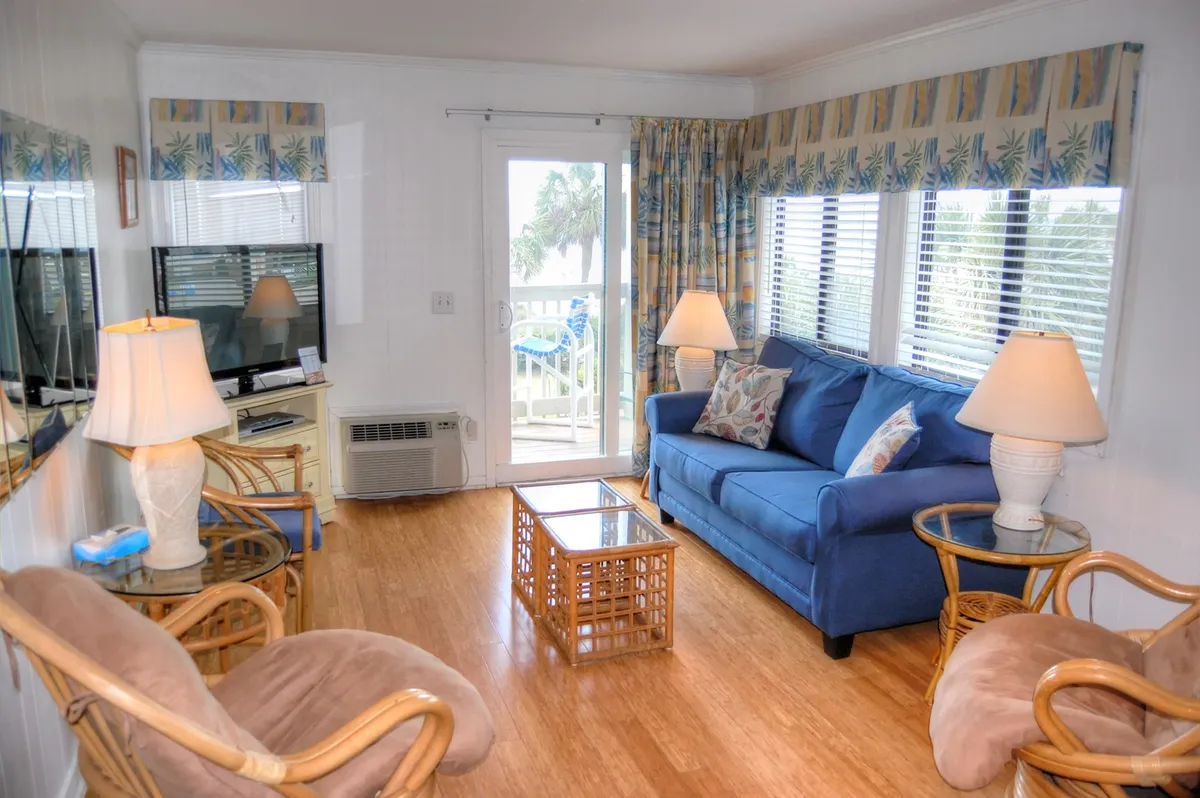 This image showcases a comfortable and stylish interior living space, featuring a blue sofa, coffee table, large windows letting in natural light, and decorative cushions. The scene feels bright and welcoming, suggesting a convivial area adjacent to the kitchen.