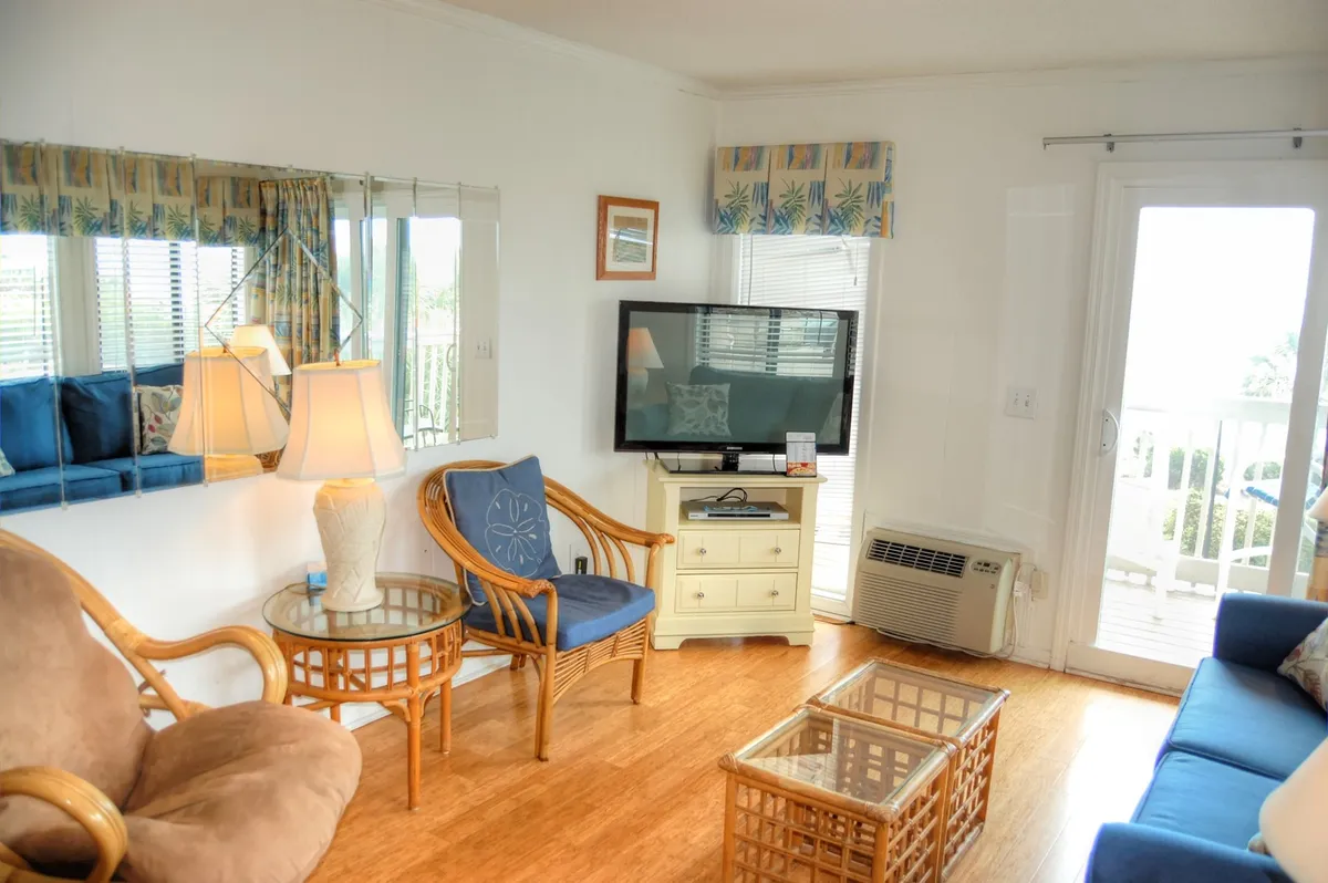 A comfortable interior lounge area featuring a blue sofa, glass coffee table, TV, and rattan chairs. The space is welcoming and practical, ideal for relaxing after a day at the pool or participating in on-site activities. The design emphasizes comfort and homeliness for extended stays.