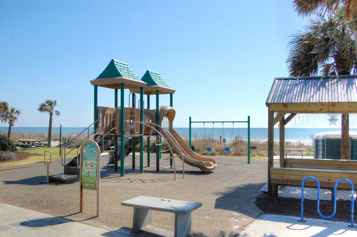 An inviting playground structure and swings set against an oceanfront backdrop, with picnic tables and palm trees nearby. Families and children can be seen enjoying playtime and casual picnics, with the blue sea stretching out beyond.