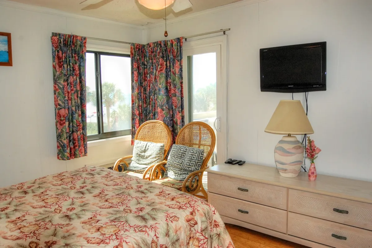A tranquil interior featuring a floral-patterned bedspread, wicker chairs, a window with curtains, and a TV mounted on the wall. The space exudes relaxation and comfort, inviting guests to enjoy cozy mornings or watch the sunset from inside.