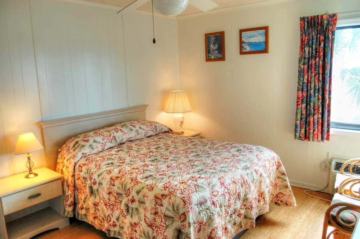 This image shows a tranquil bedroom with a bed featuring floral bedding, soft lampshade lighting, curtains with a floral pattern, and wall art. The scene is cozy and inviting, suggesting a restful space perfect for unwinding after a day at the beach.