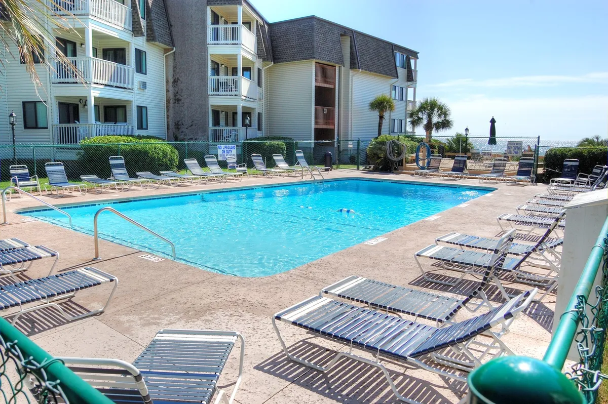 A bright and lively swimming pool scene featuring lounge chairs, surrounding greenery, and the balconies of the residential building. The setup encourages both sunbathing and socializing, with the building's proximity making it clear this is a core amenity for residents and guests.
